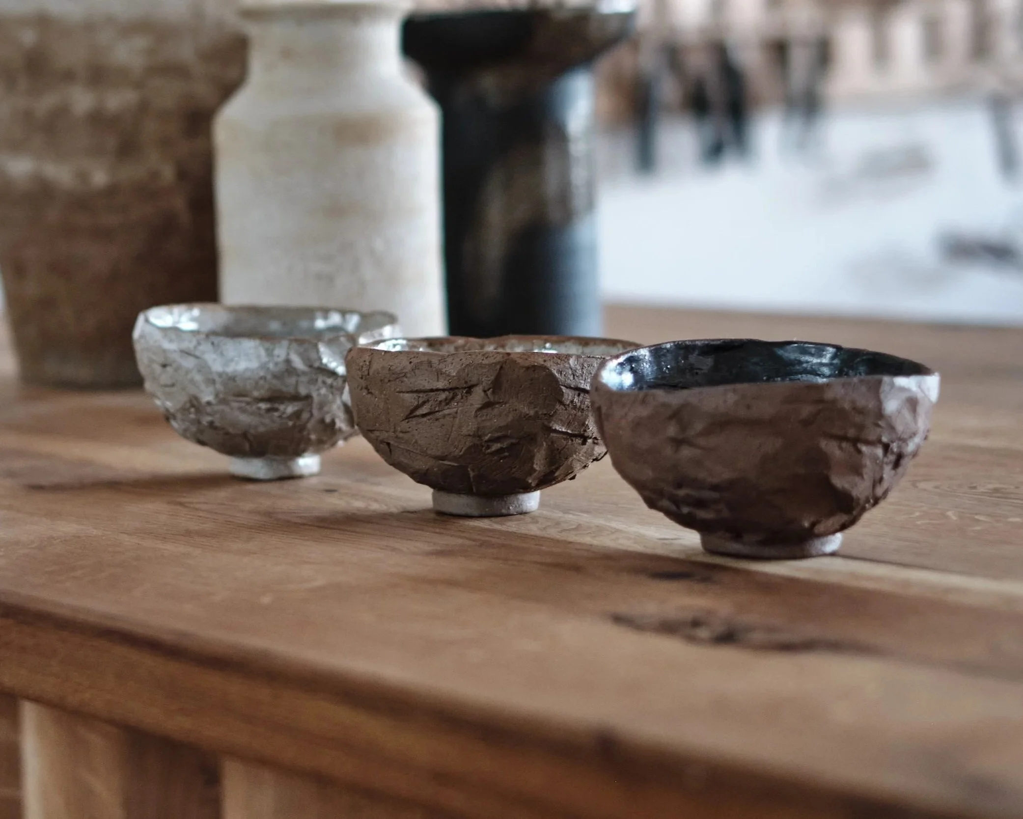 Vier kleine Schälchen aus verschiedenen Materialien auf einem Holztisch, im Hintergrund ein Milchflasche und ein geschwungener Gegenstand, möglicherweise ein weiterer Behälter.