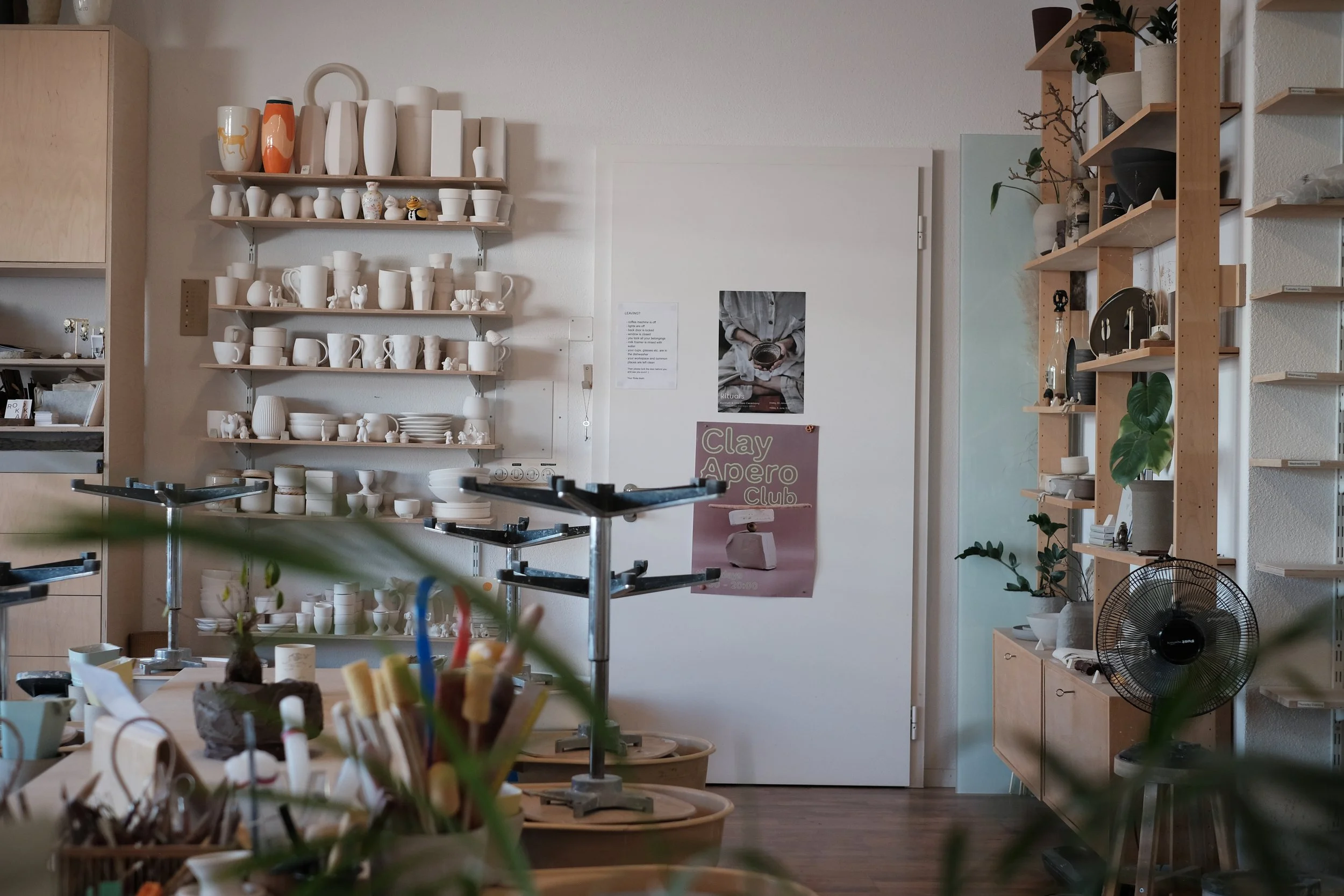 Ein Raum mit weißen und natürlichen Holzmöbeln, Regalen voller Keramikvasen, Tassen und Dekorationsgegenständen. Es gibt Pflanzen und Poster an der Wand, eine elektrische Heizung auf dem Boden, und in der Mitte befindet sich eine Arbeitsfläche mit Pf