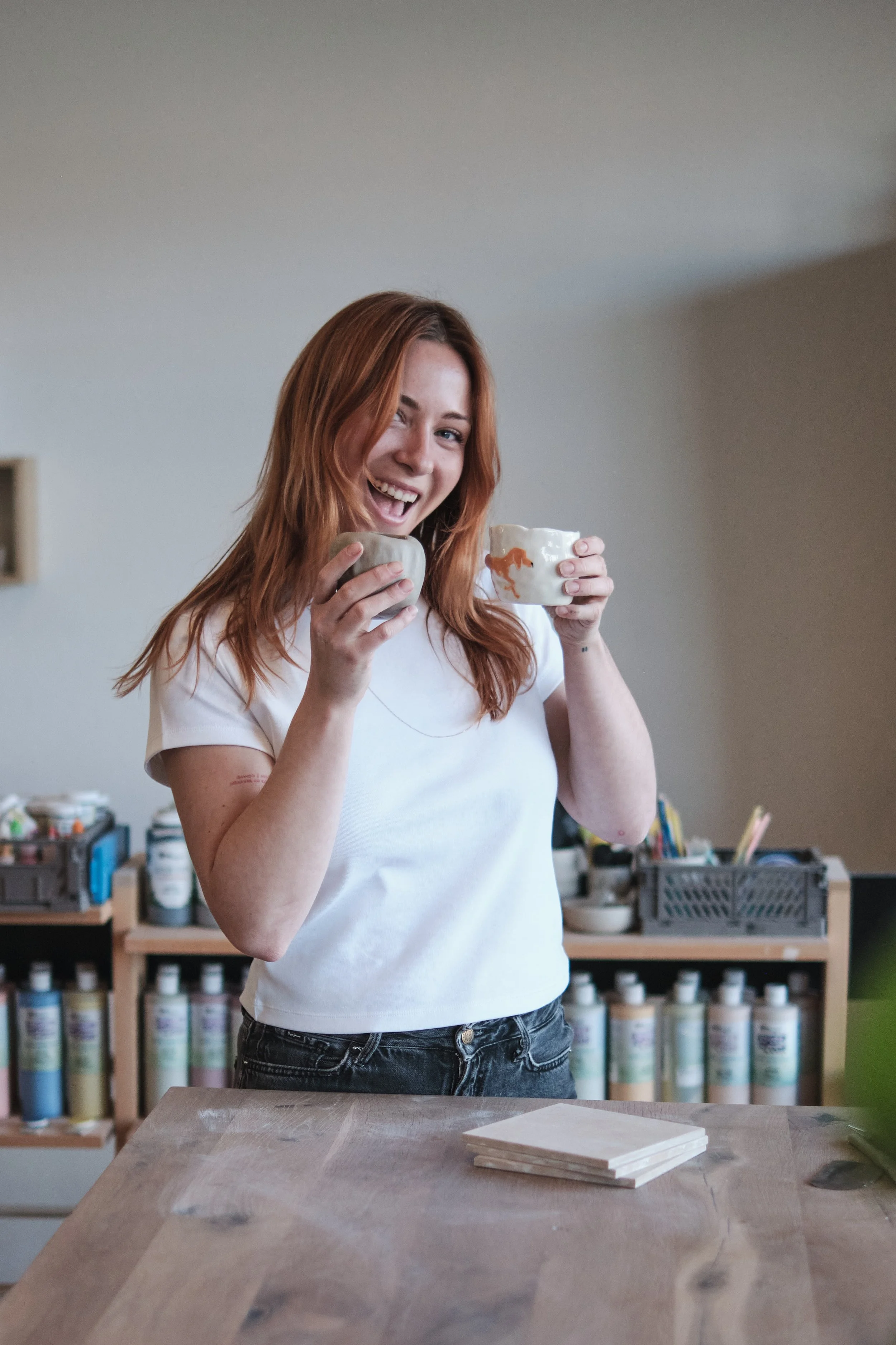 A woman with red hair writing on black tiles arranged on a wooden board, with a large green plant in the foreground and a blurred background of shelves.