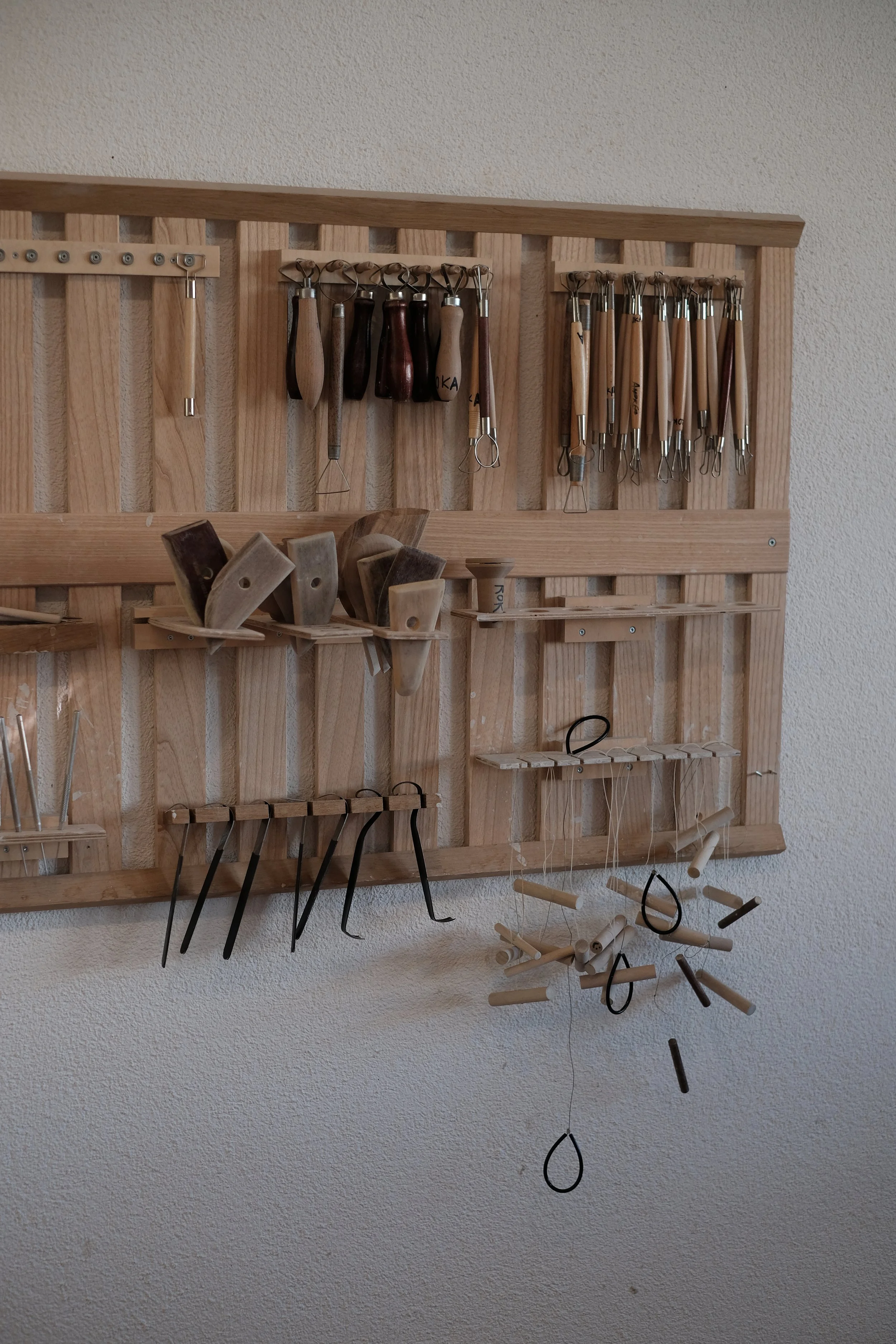 A wall-mounted wooden tool board holding various wood carving tools, including chisels, gouges, and carving knives, with some hanging from hooks and others placed on small shelves.
