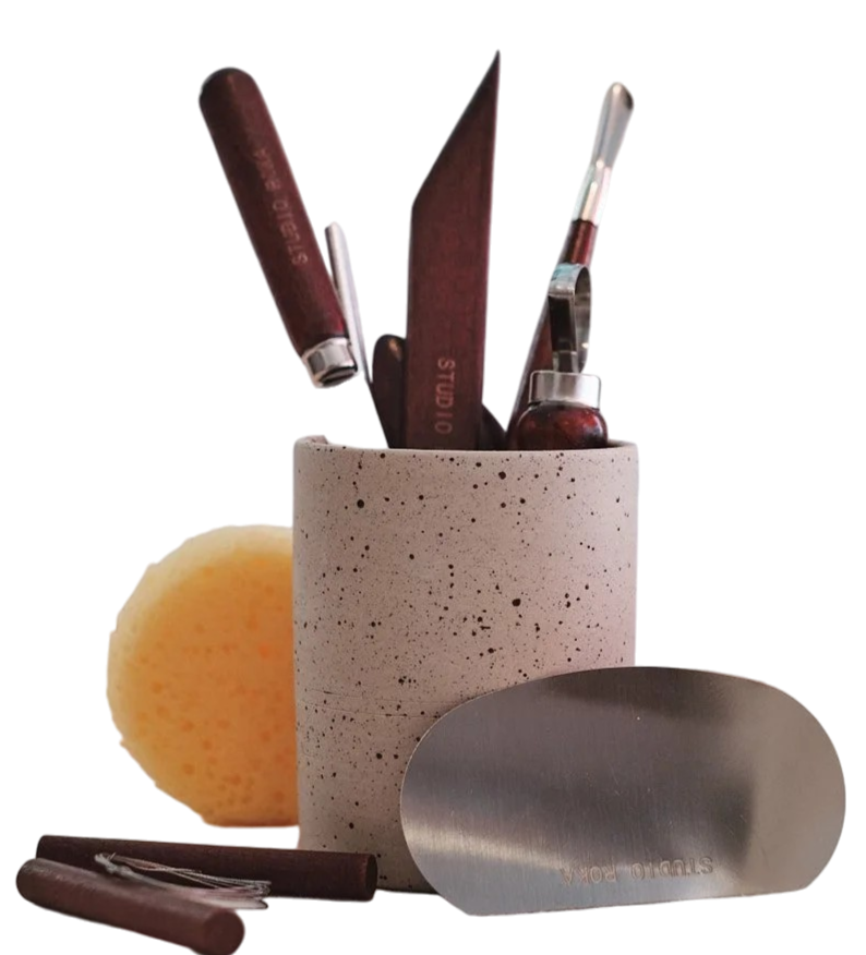 Küchenutensilien wie Messer, Teigschaber und Schneebesen in einem grauen Keramikbehälter, mit einem Stück Käse und einem Metallheber daneben.