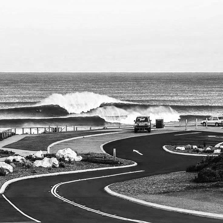 A black and white photo of a coastal road with cars parked by the side, curving toward the ocean where large waves are crashing on the shore.