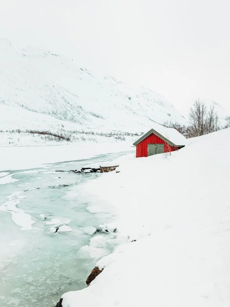 A red boathouse on snow-covered ground next to a partially frozen river in a snowy mountain landscape.