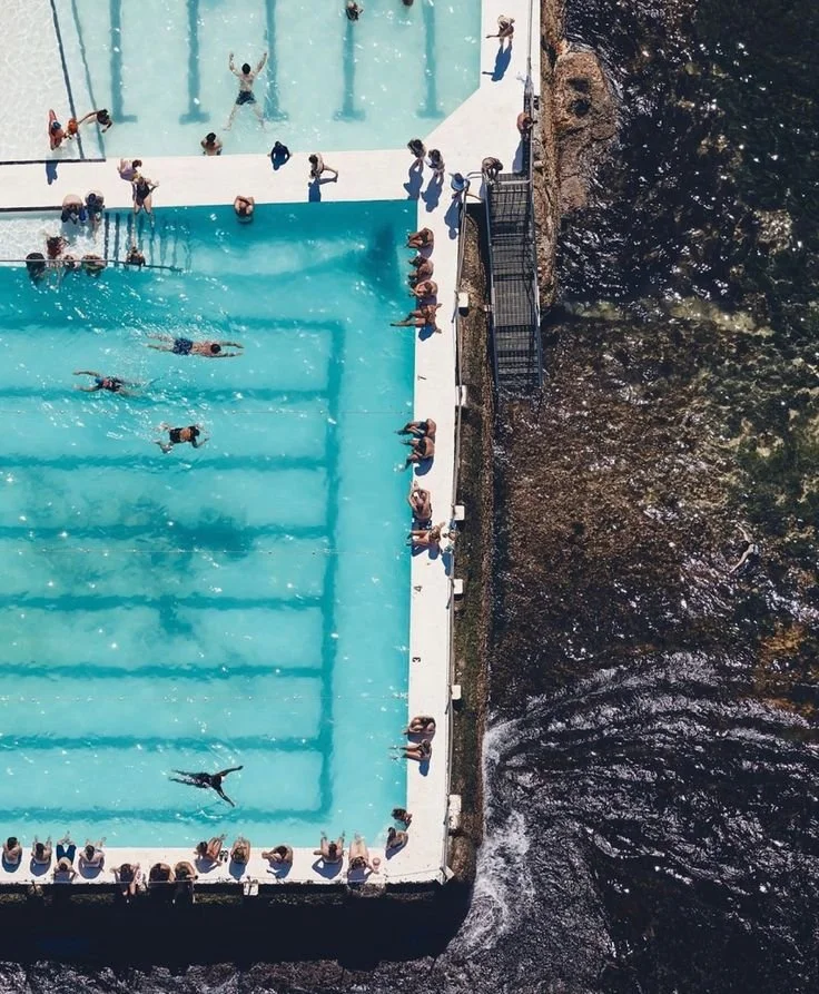 An aerial view of a swimming pool with people swimming and lounging around, next to a river or stream.