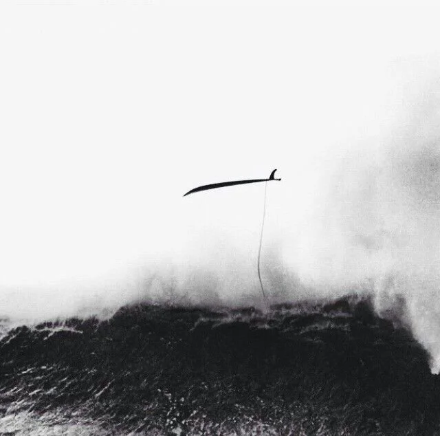 A person appears to be underwater with a large wave crashing above, with a thin line or string hanging down, all in black and white.