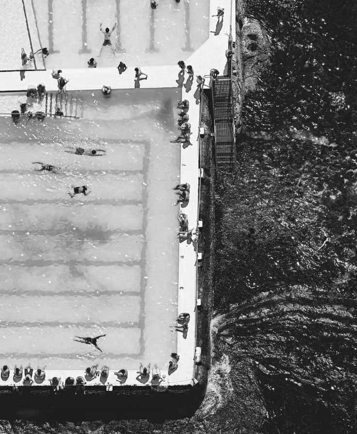 Bird's eye view of a busy public swimming pool with people swimming, sunbathing, and socializing around the edge, surrounded by water.