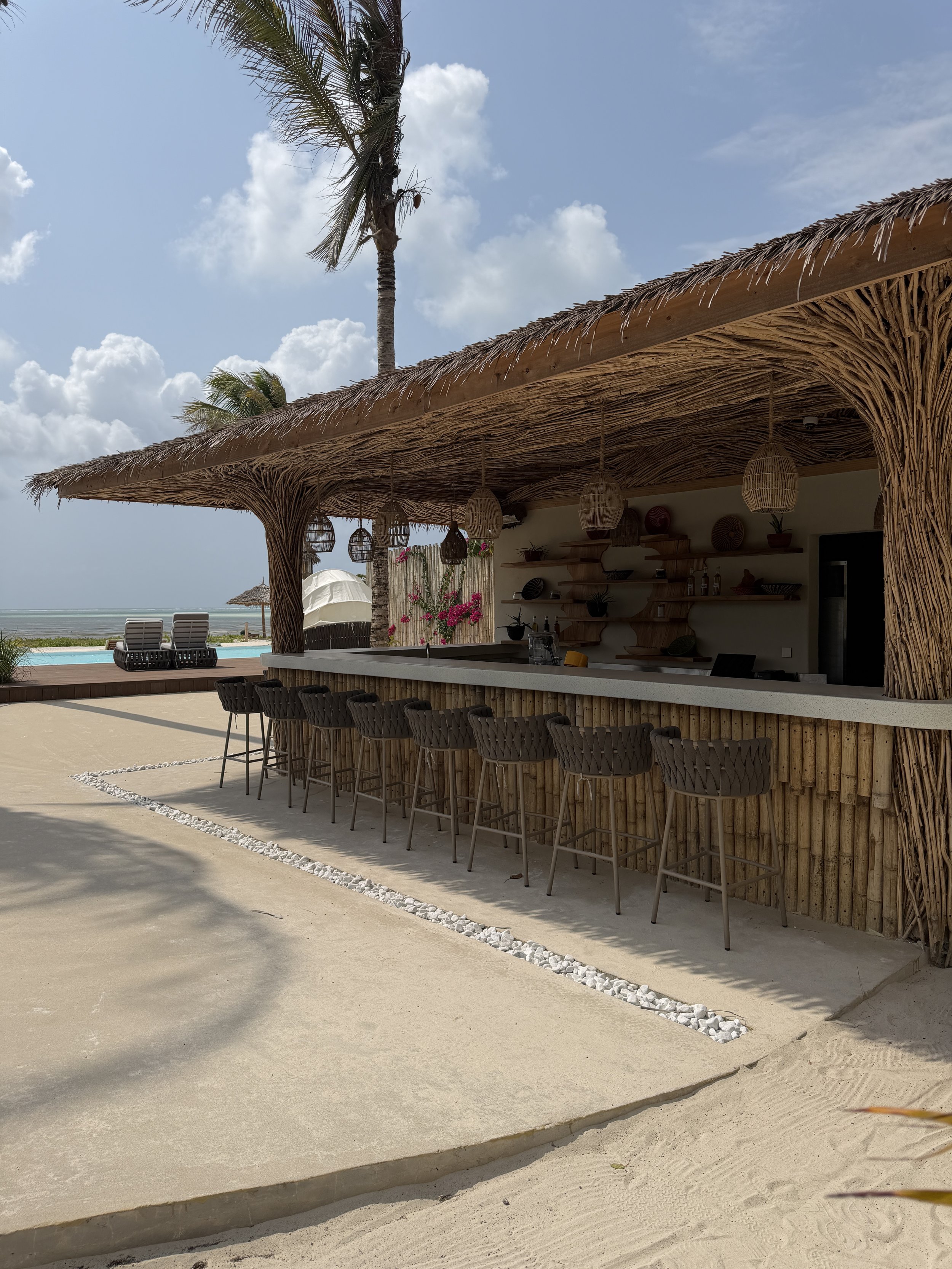 Sfeervolle bar van ENVI Paje op Zanzibar, met een houten bar met prachtig houtsnijwerk, rotan barkrukken en natuurlijke materialen in tropische stijl.