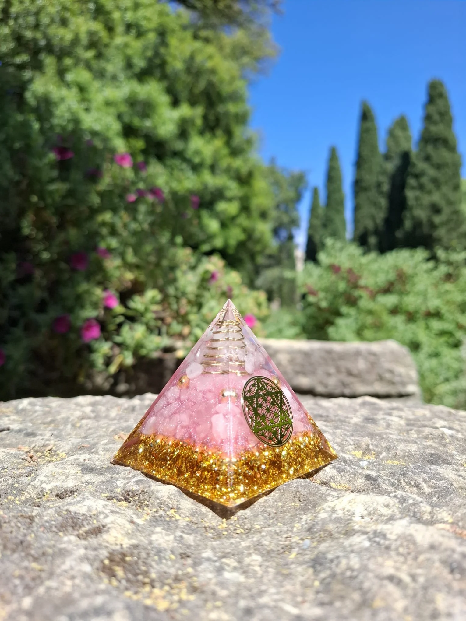 Pyramide orgonite au soleil, en cristal de roche et quartz rose, entourée de plantes dans un jardin zen