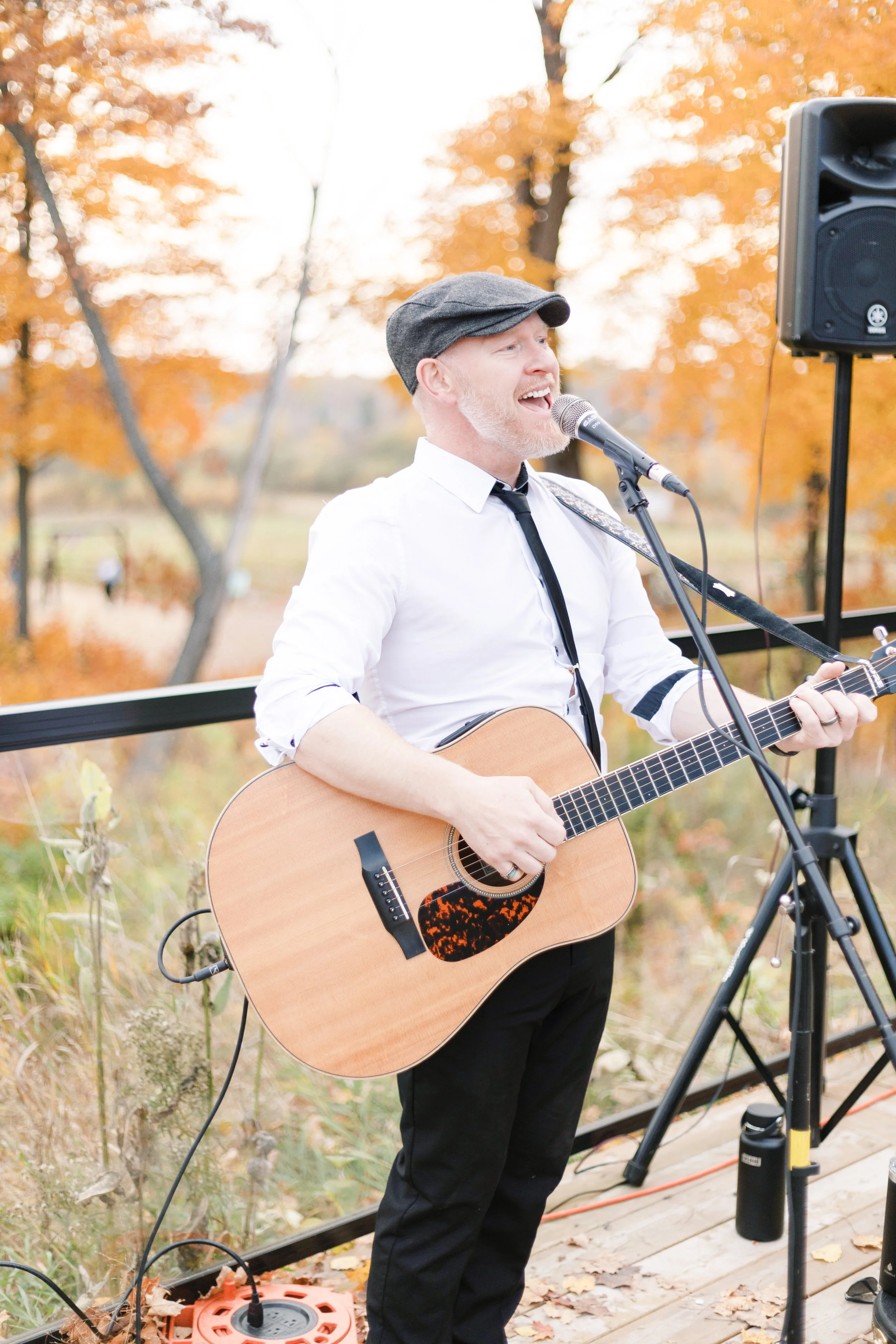 A man singing and playing an acoustic guitar outdoors with autumn-colored trees in the background.