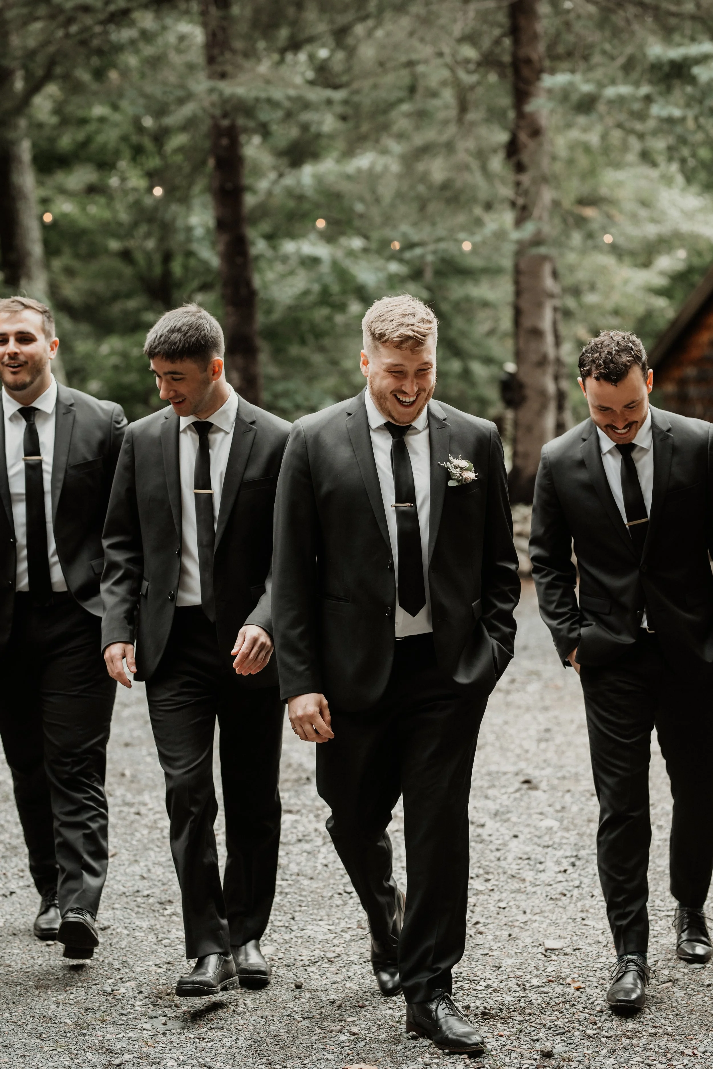A group of five men dressed in black suits, white shirts, and black ties walking outdoors on a gravel path in a wooded area, smiling and laughing together.