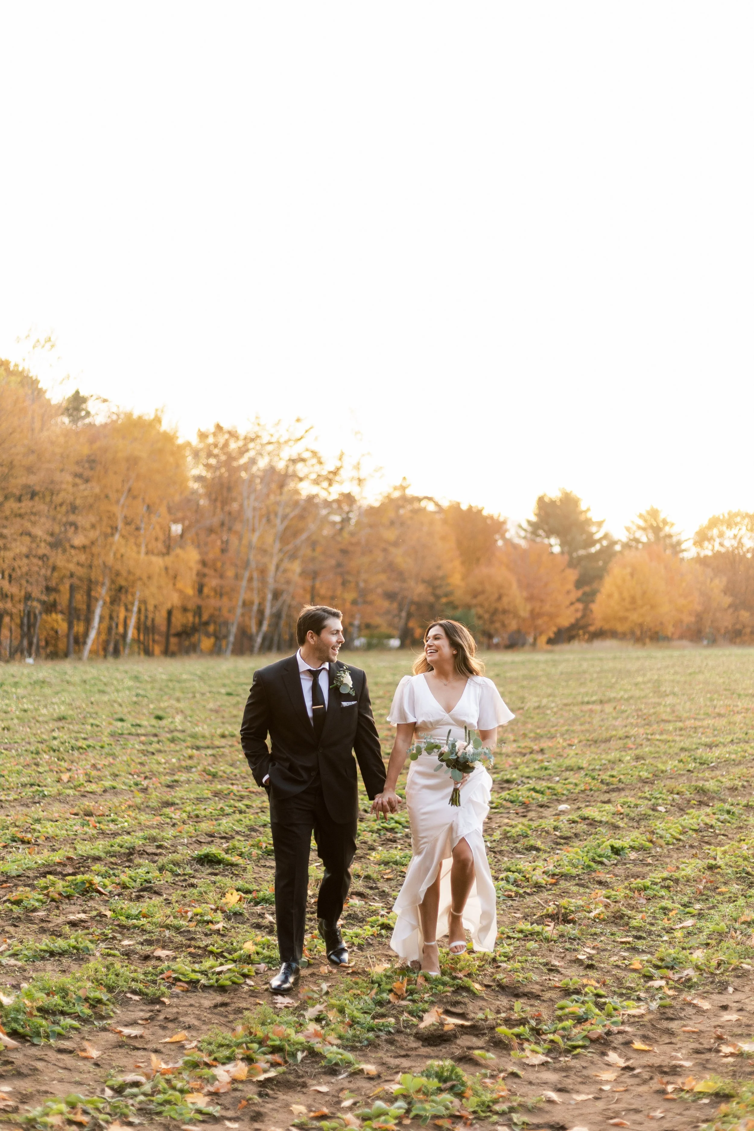 A bride and groom walking hand in hand through a field during sunset, surrounded by trees with autumn foliage.