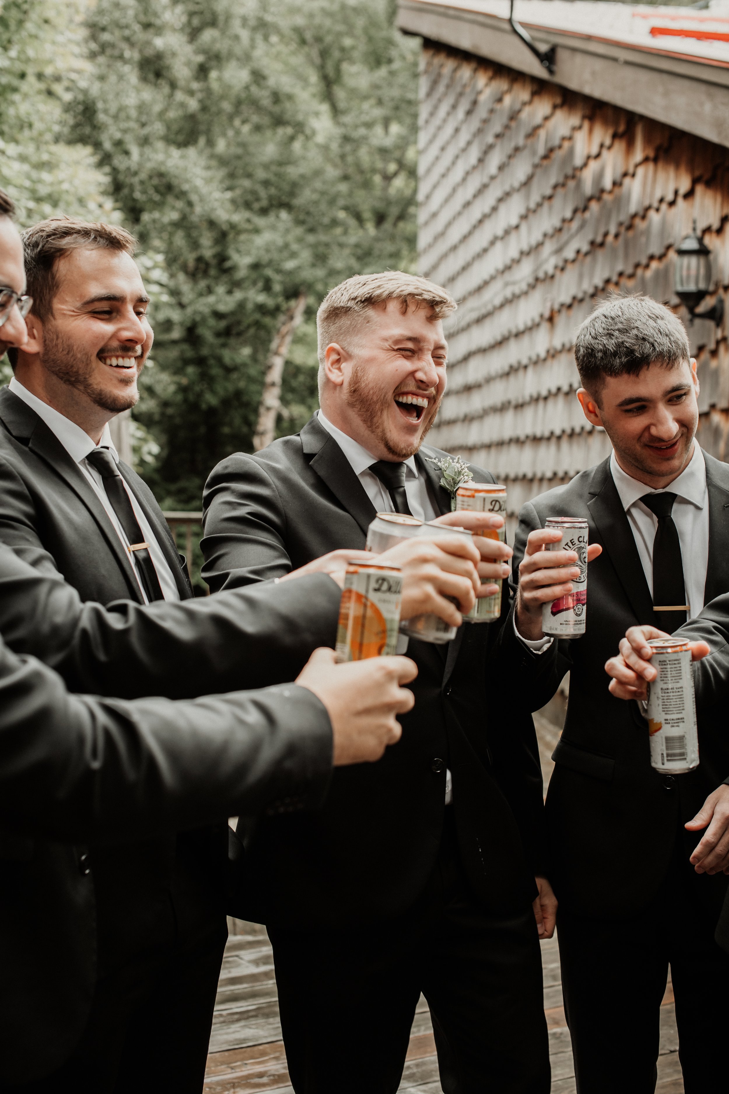 Group of men in suits smiling and laughing while holding canned beverages outdoors