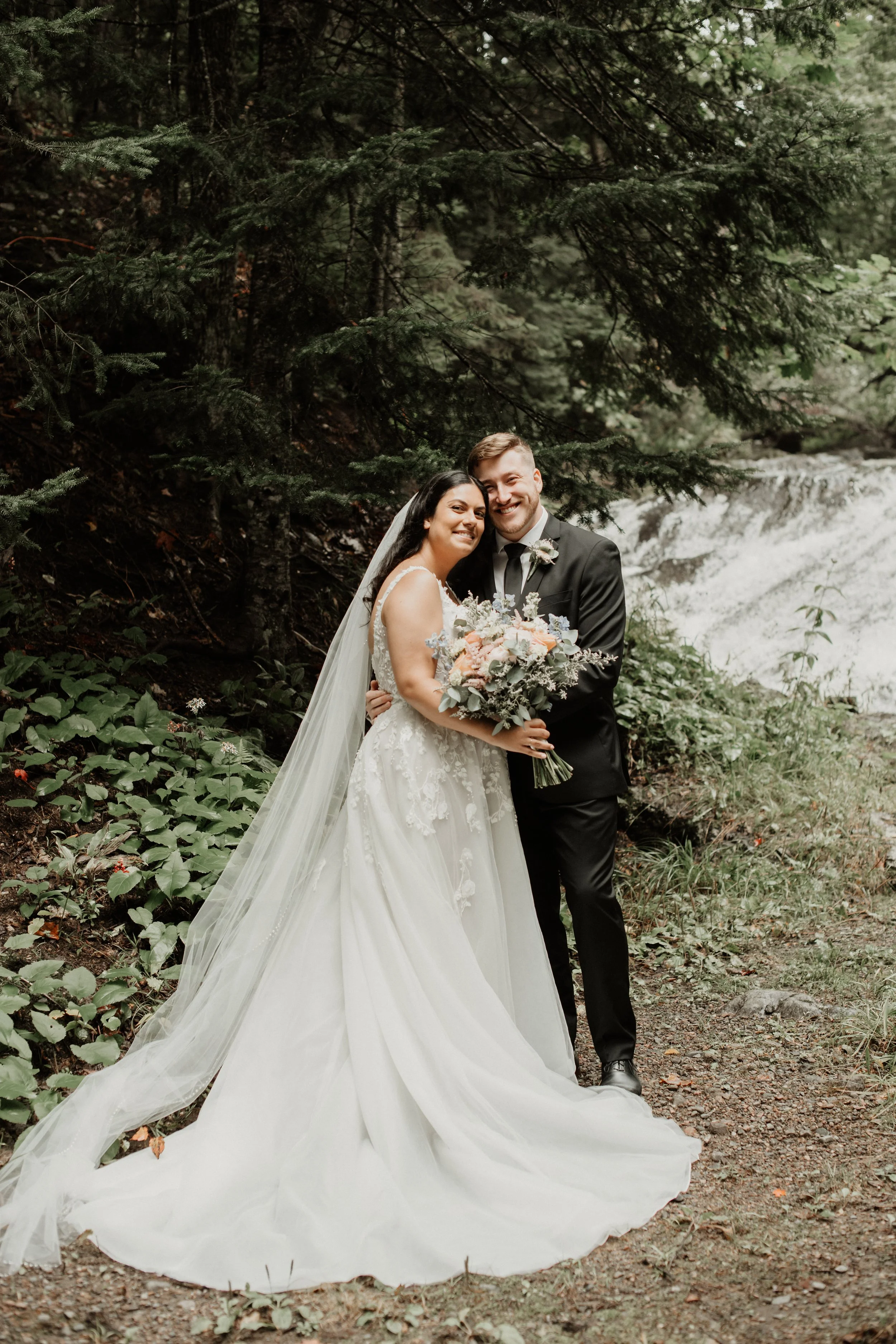 A newlywed couple in wedding attire standing outdoors in a forest near a waterfall, smiling and holding a bouquet of flowers.