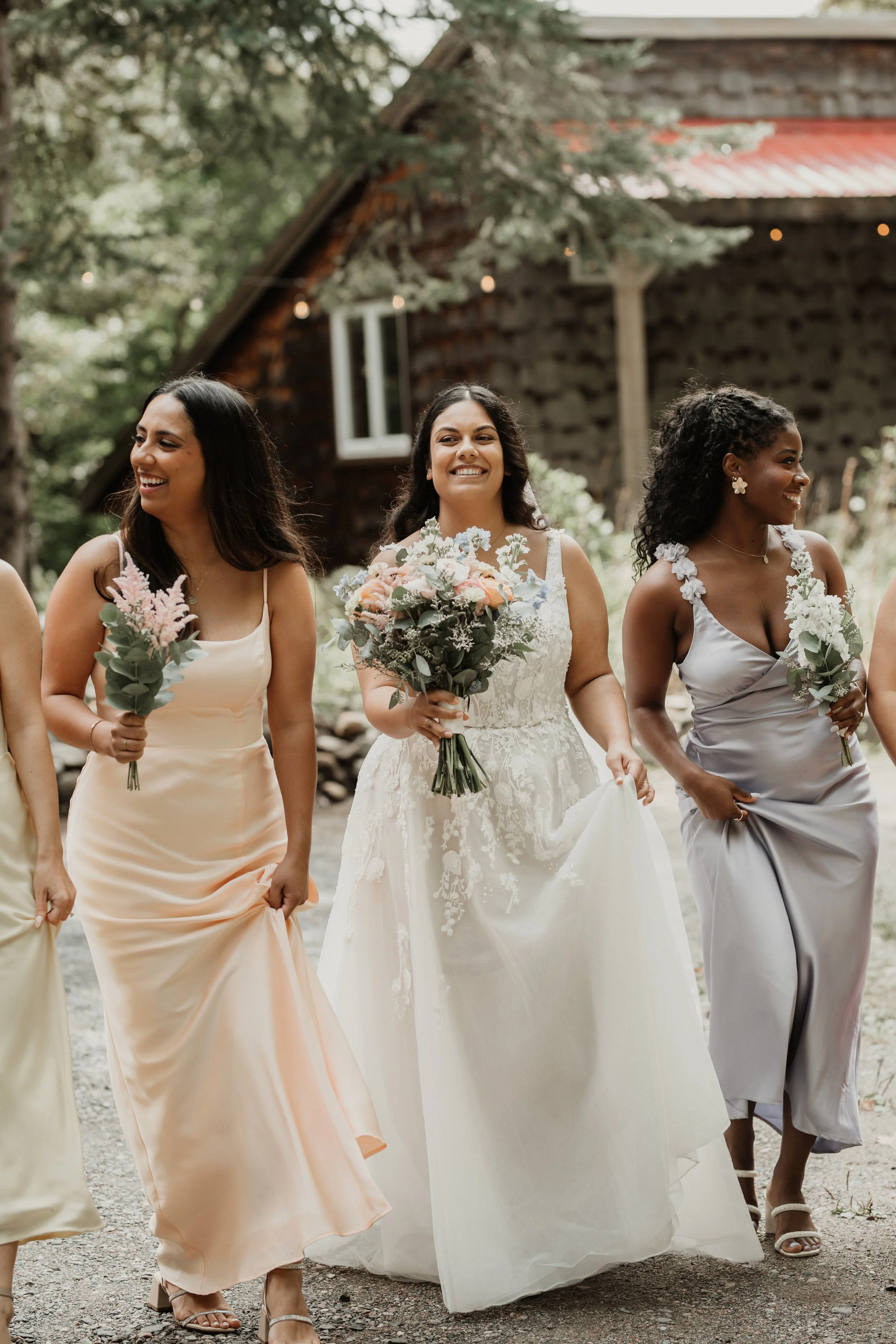 Group of women in formal dresses holding bouquets, walking outdoors near a rustic wooden building with trees in the background, during a wedding or special event.