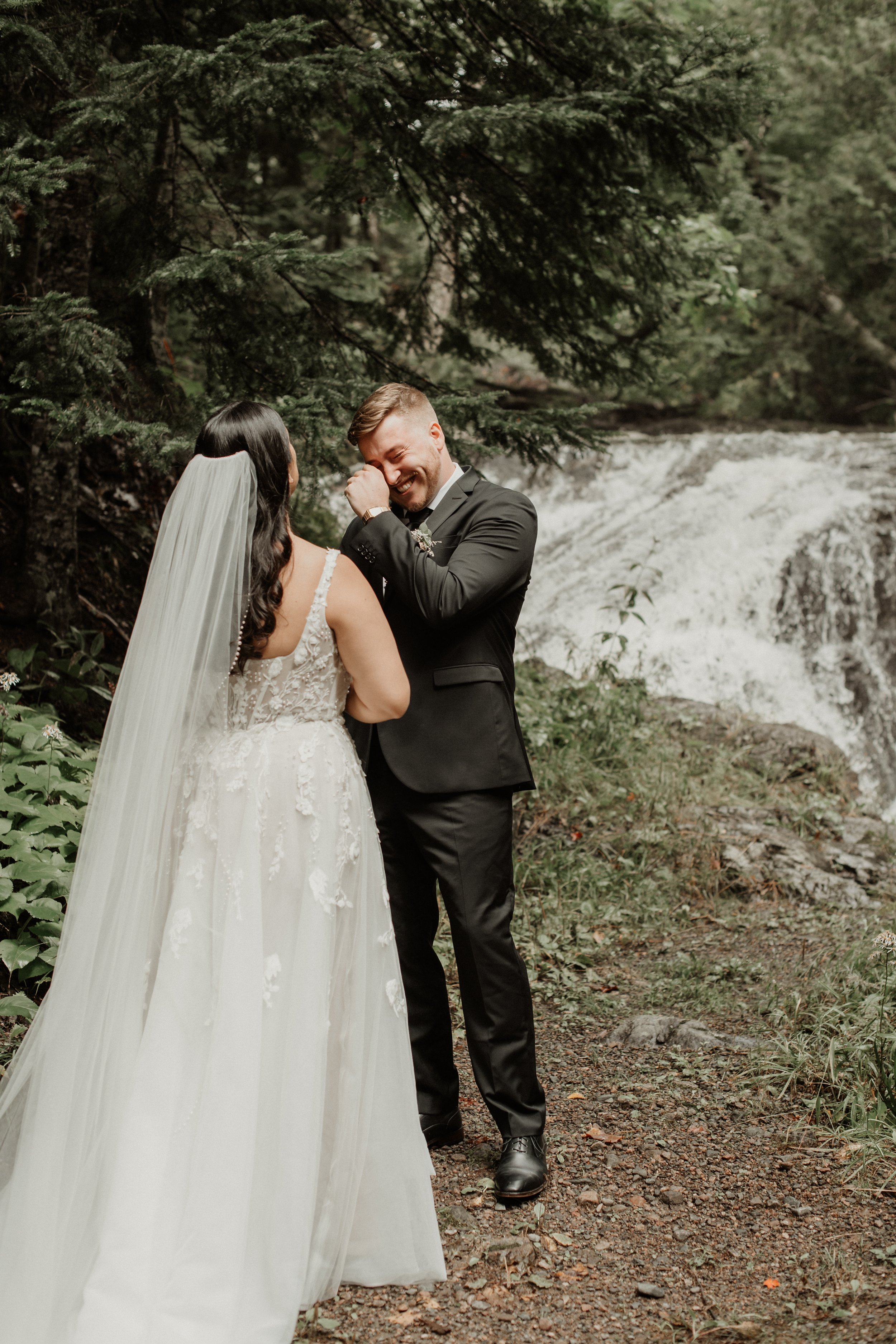 A bride and groom standing outdoors near a river with a waterfall in the background, smiling and sharing a moment of joy.