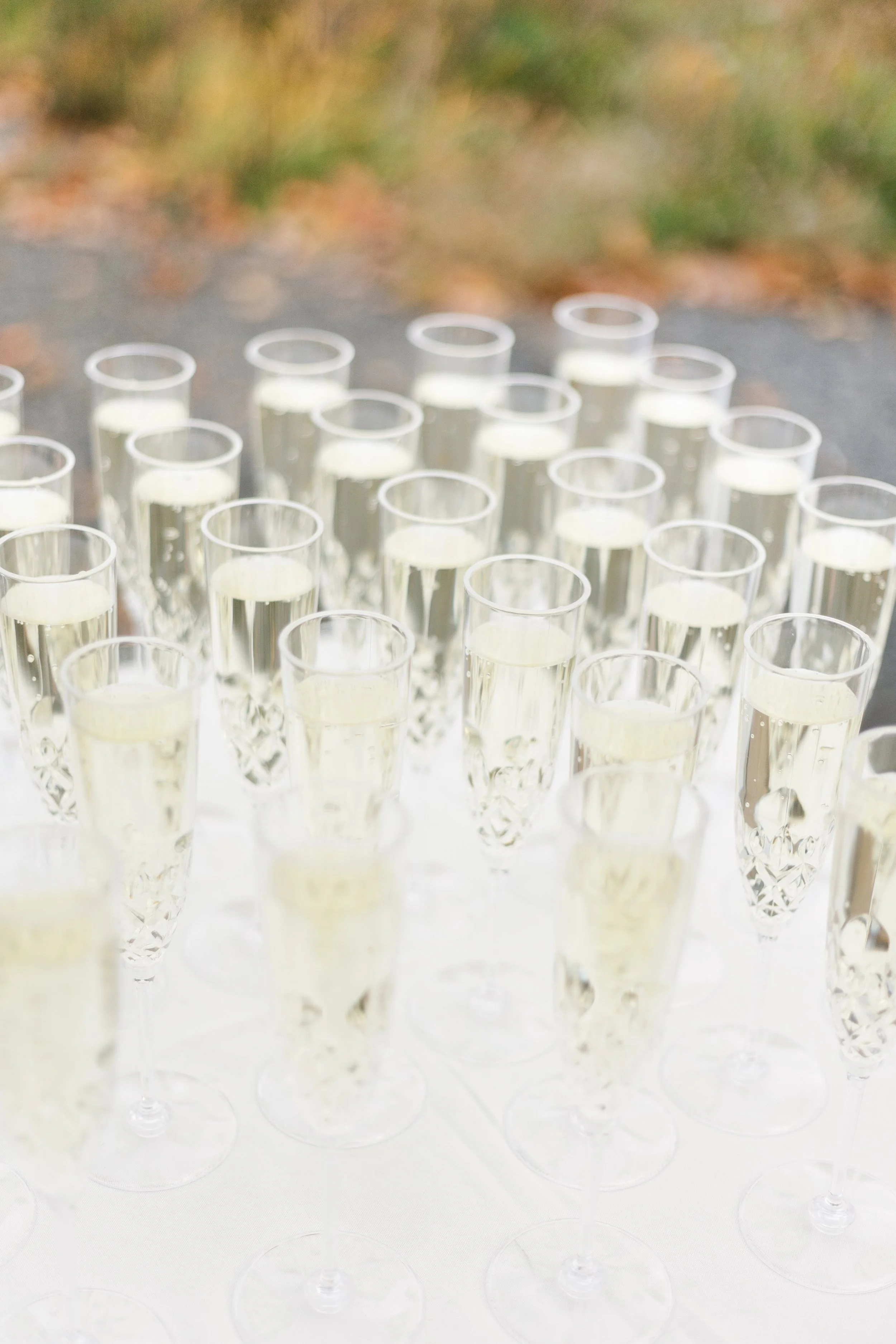 Multiple clear champagne flutes filled with champagne arranged on a white surface with a blurred outdoor background.