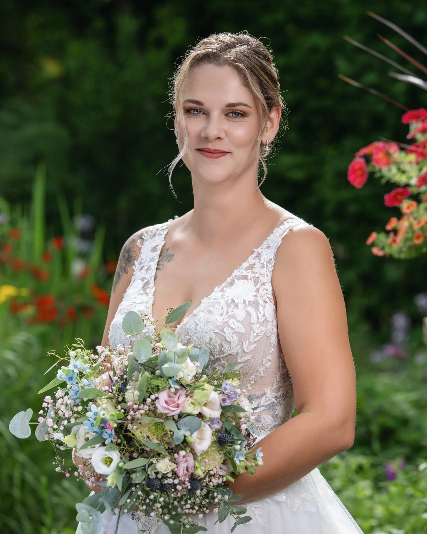 Eine junge Frau in einem weißen Hochzeitkleid hält einen Blumenstrauß vor einem grünen Garten. Sie lächelt leicht und trägt Ohrringe.