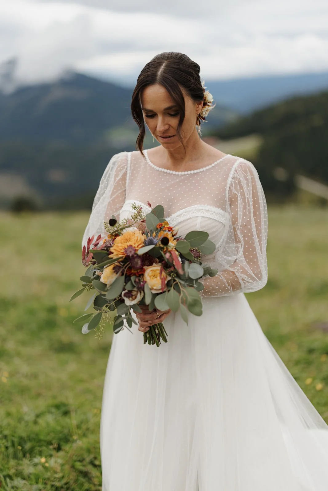 Braut in weißem Brautkleid mit Schleier hält einen bunten Blumenstrauß in einem Grünen Feld mit Bergen im Hintergrund.