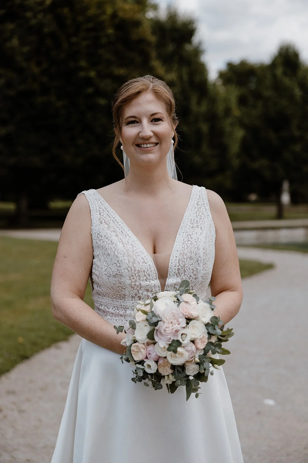 Junge Braut mit braunen Haaren, die ein weißes Hochzeitskleid trägt, hält einen Blumenstrauß aus weißen und hellrosanen Blumen in einer Parkumgebung im Freien.
