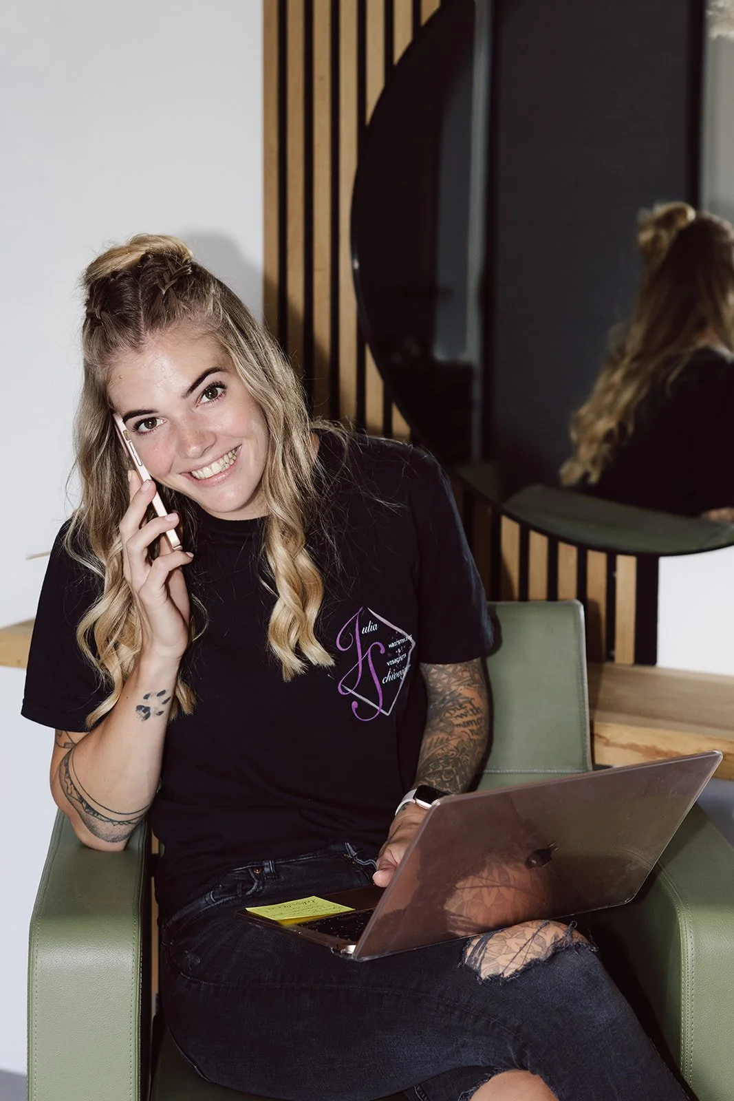 Junge Frau mit Tattoos spricht lächelnd am Telefon, sitzt auf einem grünen Stuhl mit Laptop auf dem Schoß, trägt ein schwarzes T-Shirt mit einem Logo, im Hintergrund eine große runde Spiegelung