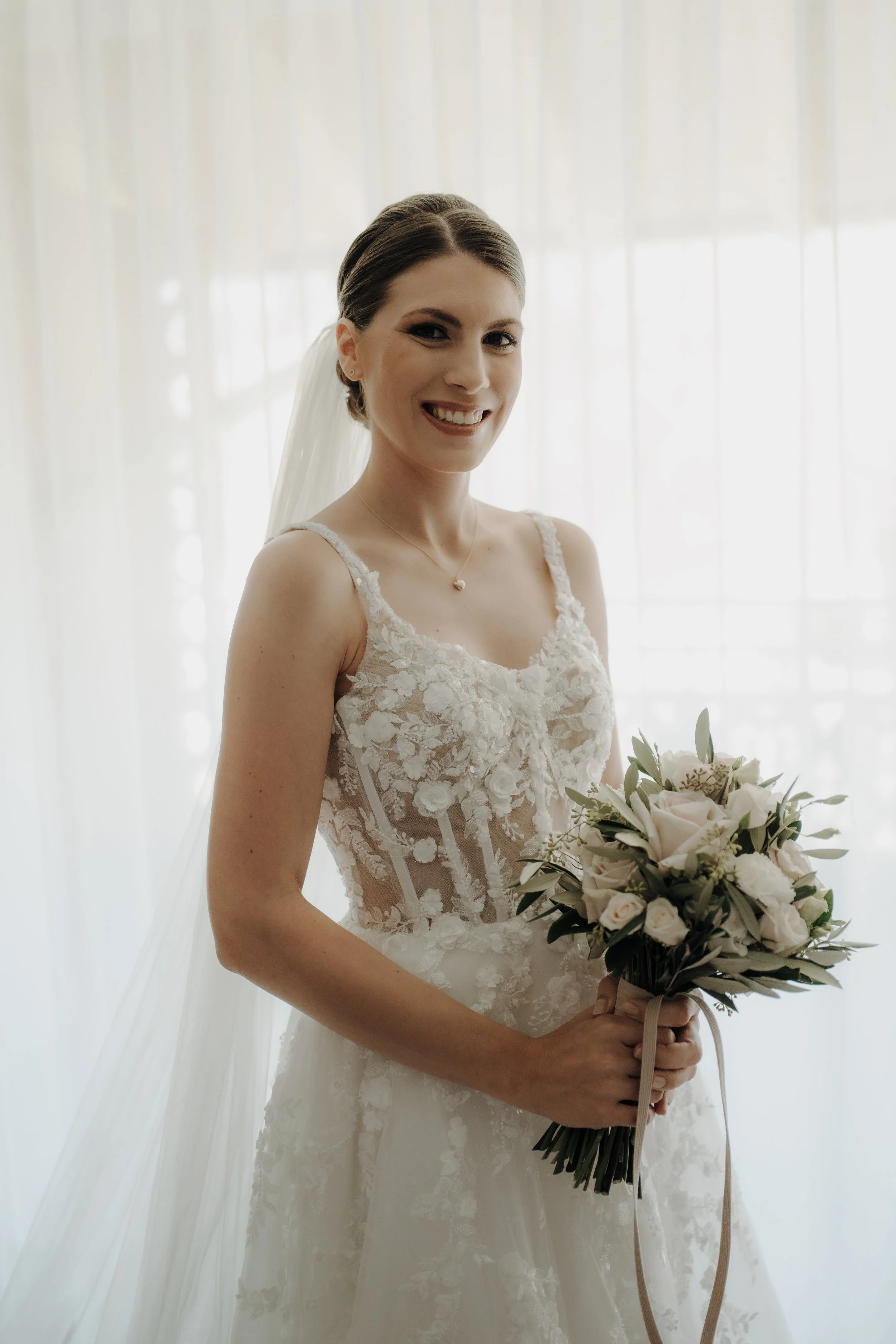 Eine glückliche Braut in einem weißen Hochzeitskleid mit Blumenstrauß vor einem hellen, lichtdurchfluteten Vorhang.
