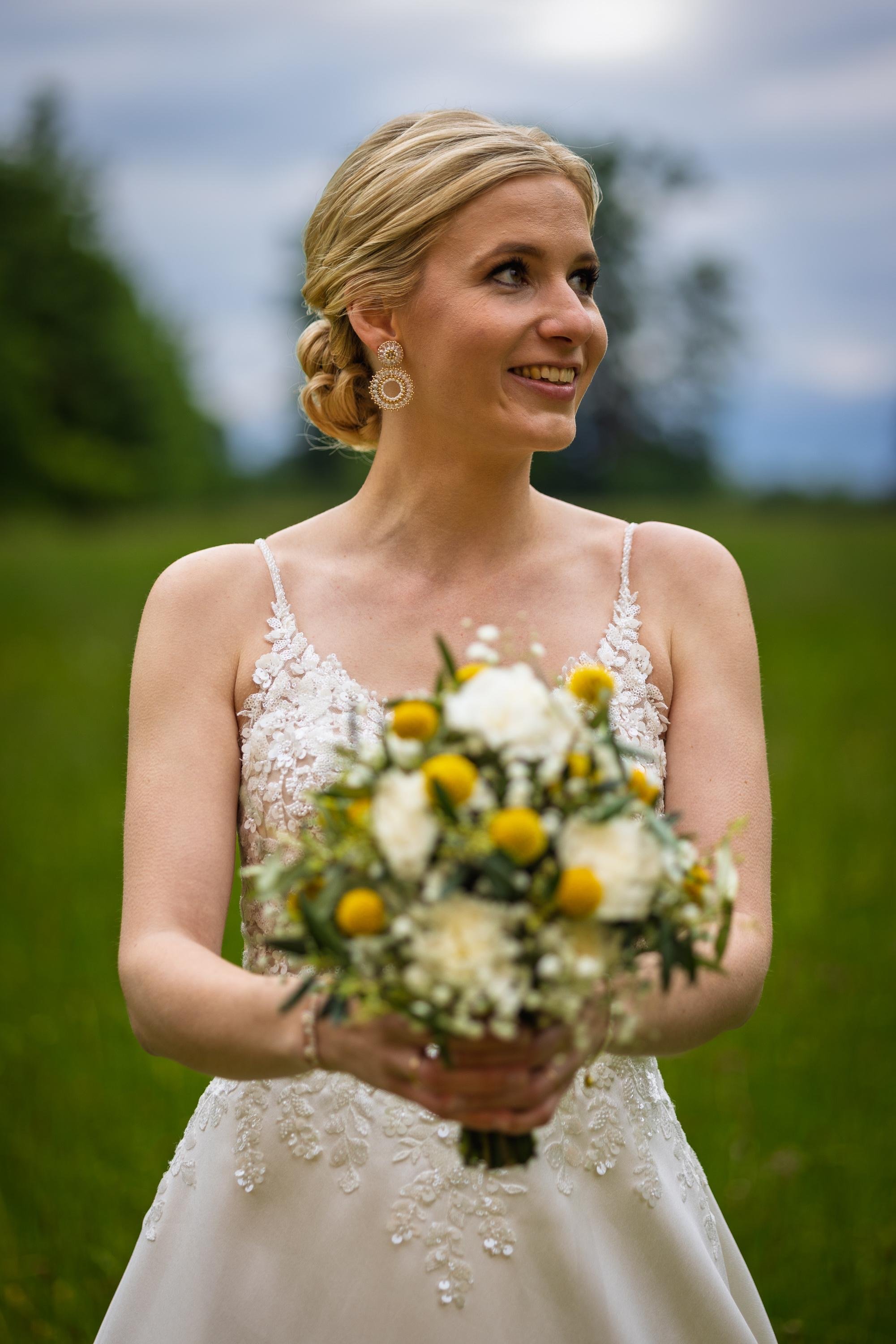Eine Frau in einem weißen Brautkleid hält einen Blumenstrauß und steht im Freien bei bewölktem Himmel.