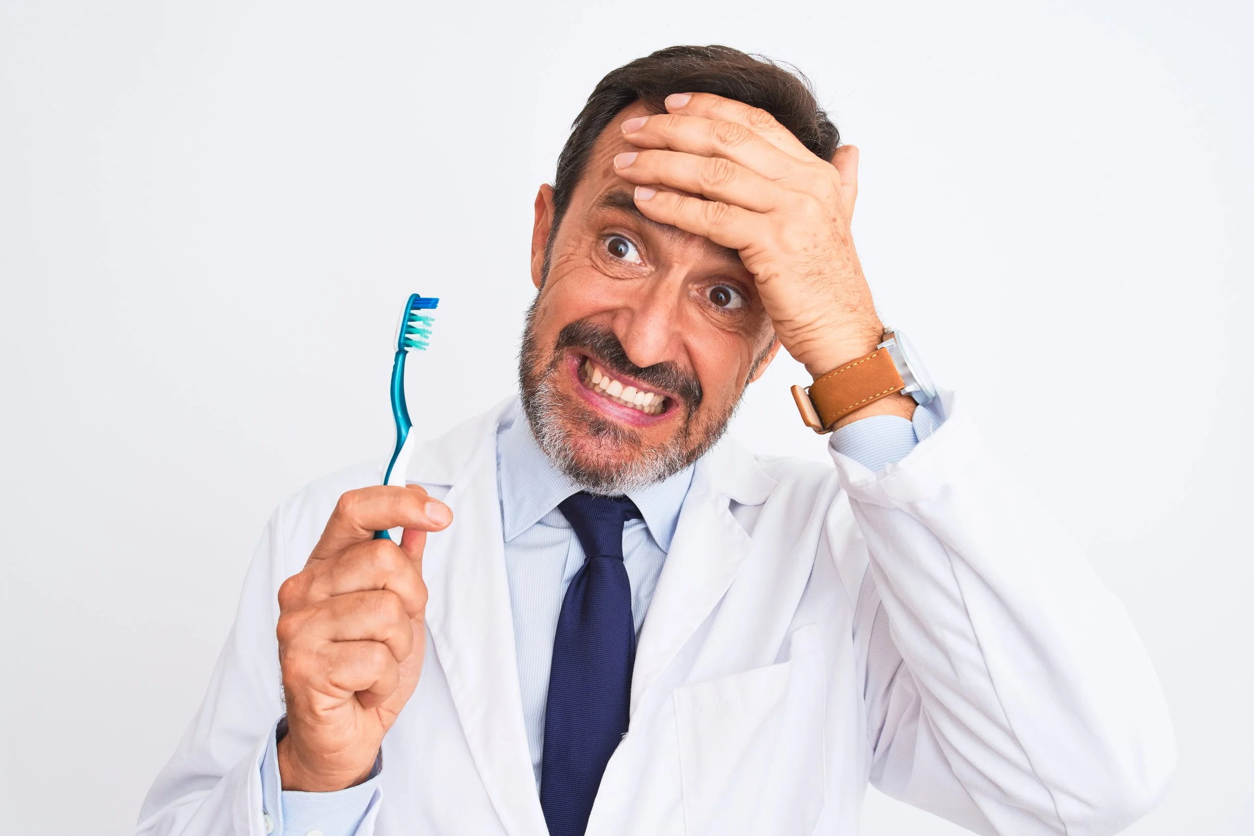 photograph of a frustrated dentist holding a hand to his forehead. the other hand is holding a toothbrush