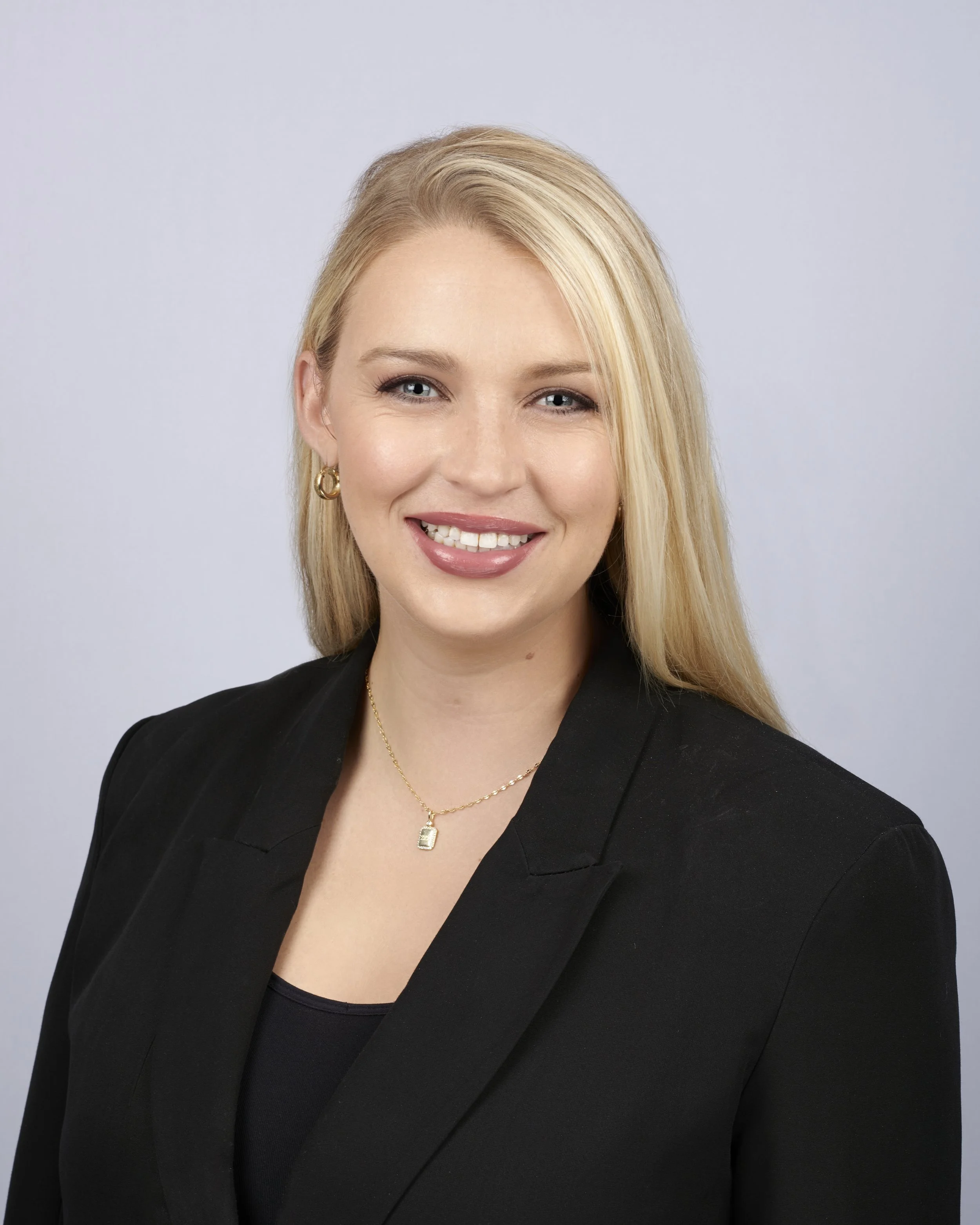 A smiling woman with long blond hair, wearing a black blazer and pearl earrings, against a white background.