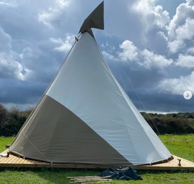 A large, triangular-shaped teepee tent set up outdoors on a grassy field with a platform base, under a cloudy sky.