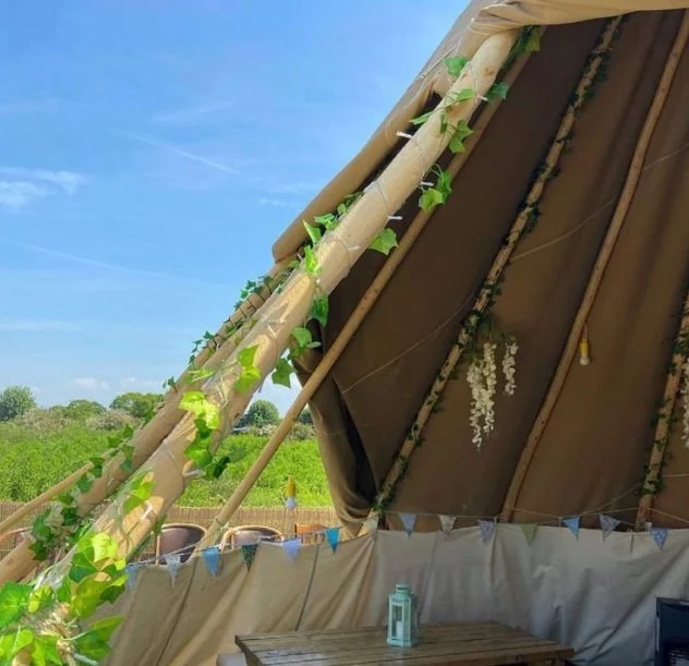The interior of a large beige canvas tent decorated with green vines and a string of colorful bunting, with a small wooden table and a mason jar inside, set in a grassy field under a blue sky.