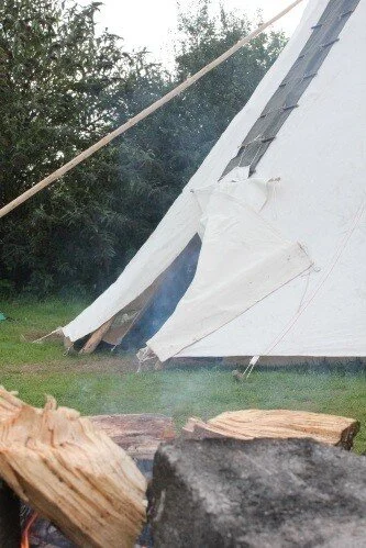 Close-up of a campfire with logs, a tent in the background, and trees surrounding the area.