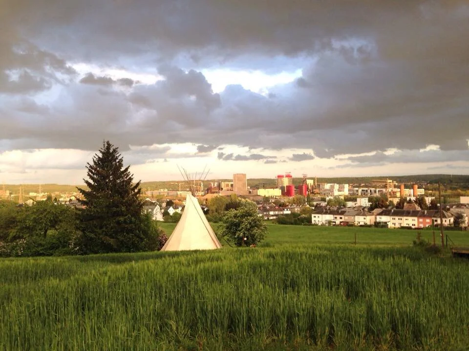 Green field with a tent and trees, with a city skyline and cloudy sky in the background.