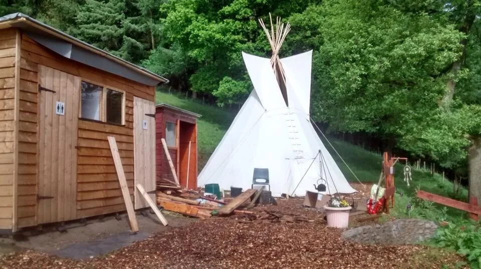 A small outdoor area with a wooden shed, a small red shed, and a white teepees tent. Surrounding trees and a wooden fence, with various gardening and outdoor items scattered around.