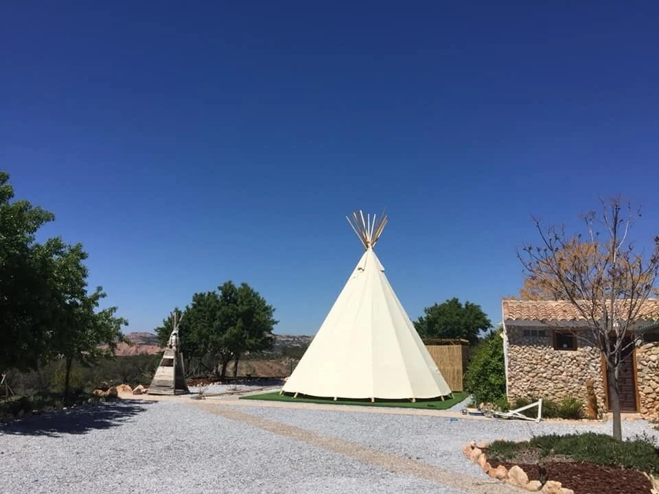 A large white teepee tent with wooden poles on top, set on a gravel path in a sunny outdoor area. There are trees and a stone building nearby, with a clear blue sky overhead.