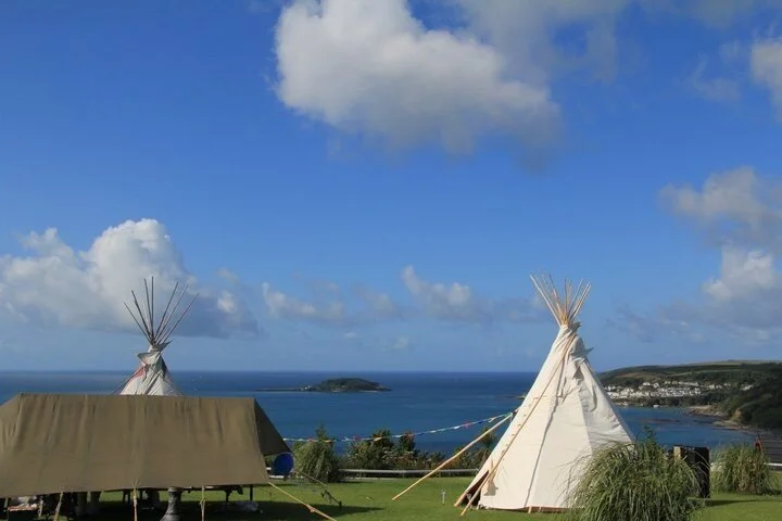 Three tents, two tipi-style and one rectangular with a brown cover, on a grassy area overlooking the ocean with a small island and hilly coastline in the distance