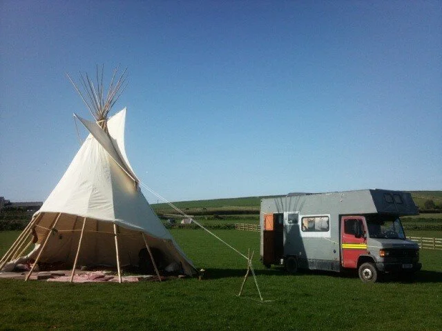 A small teepee tent set up on grass next to a parked camper van, with open fields and blue sky in the background.