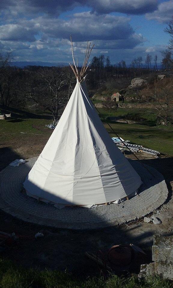 A small white teepee tent with wooden poles, set on paved ground amid a natural landscape with trees and rocks.