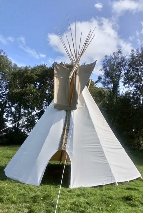 A white canvas teepee tent set up outdoors on a grassy area, with a blue sky and trees in the background.