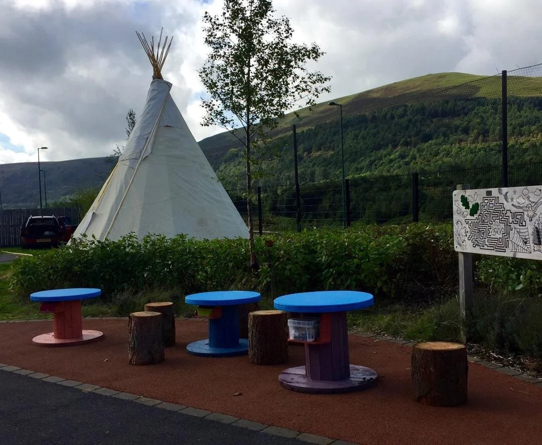 Children's outdoor play area with colorful round tables and logs for seating, a small teepee tent, and a maze on a sign, with green hills and cloudy sky in the background.