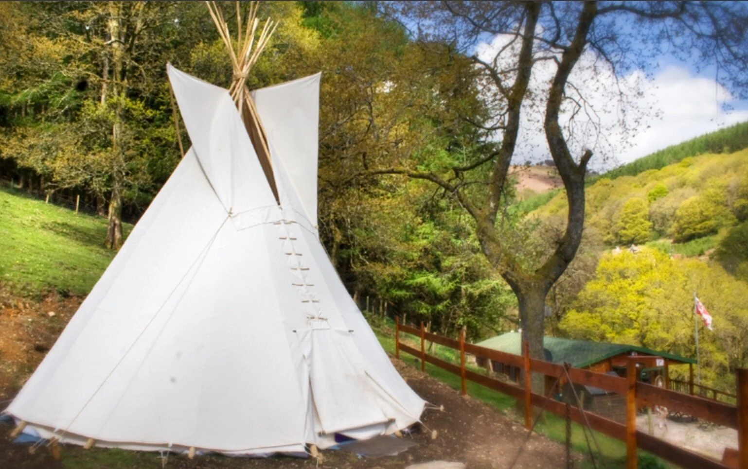 A white teepee tent set up on a grassy hillside near a wooden fence, with green and yellow trees and a small building with a green roof and an American flag in the background.
