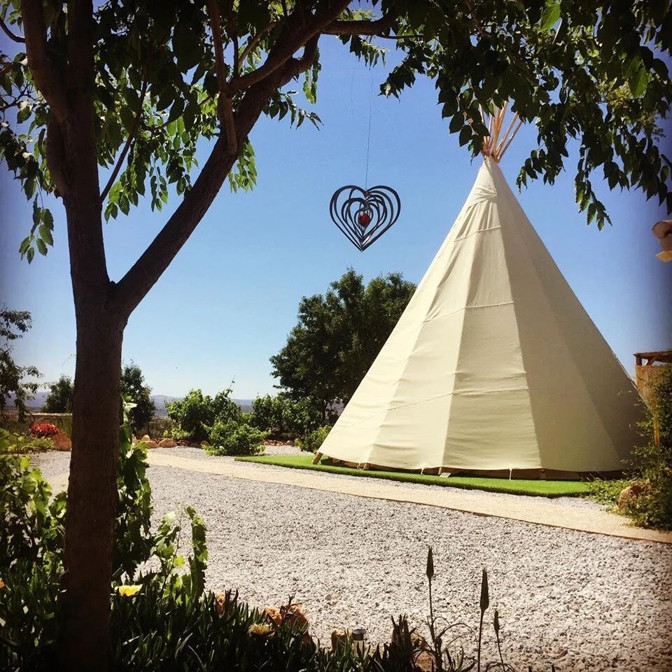 A white tipi tent set up in a landscaped area with trees and bushes, under a clear blue sky, with a black heart-shaped wind chime hanging from a tree branch.