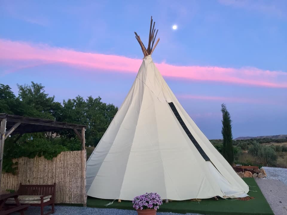 A white tipi tent with wooden poles at the top, set outdoors during dusk with a pink and purple sky and the moon visible. There is a wooden structure with a bench and a potted plant with purple flowers in front.