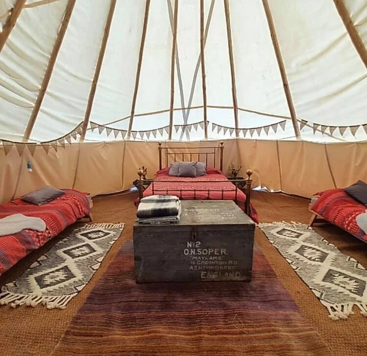 Interior of a large, beige tipi or yurt with wooden support beams, featuring a central bed, two sleeping mats with pillows, a wooden chest at the foot of the bed, and decorative rugs on the wooden floor.