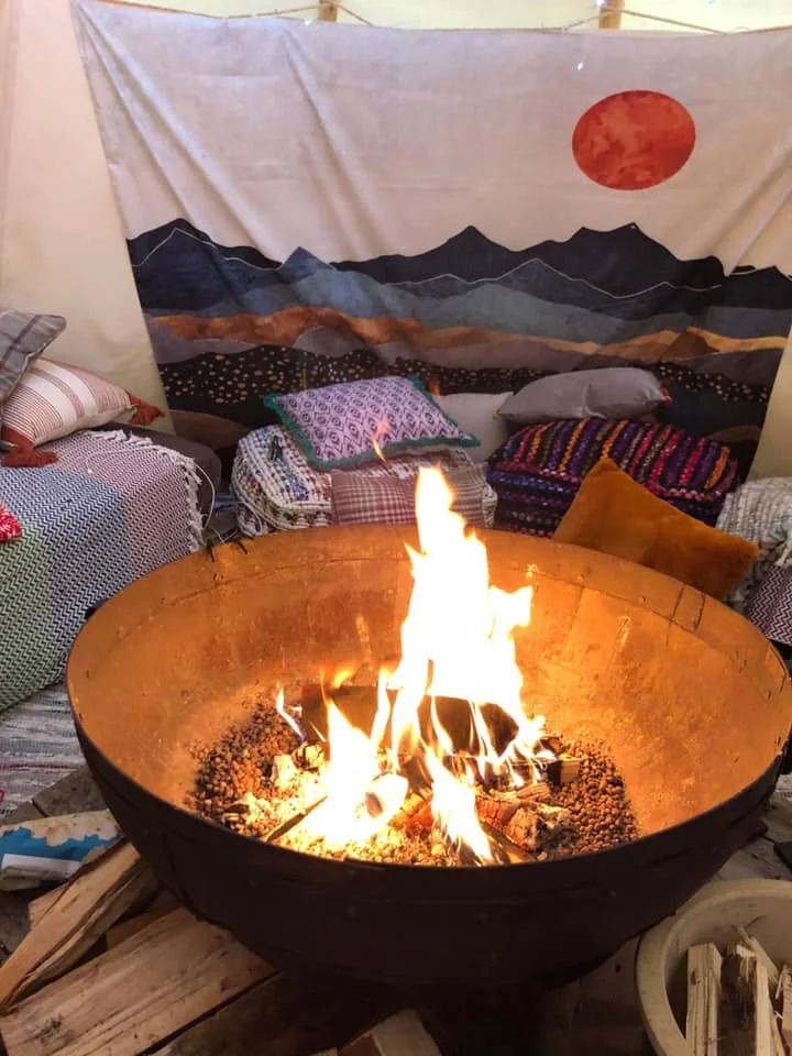 Indoor setting with a large fire in a metal fire pit, surrounded by cushions and pillows on the floor, with a tapestry depicting mountains and a red sun on the wall in the background.