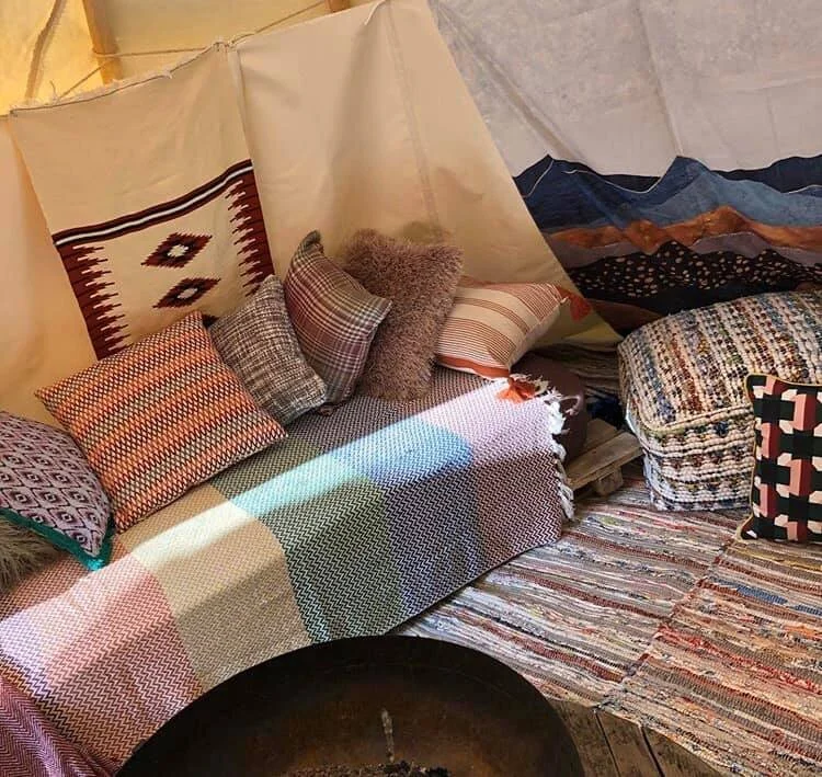 Inside a cozy room with a daybed covered in multicolored woven and patterned pillows, a patchwork blanket, and a multicolored rug, with fabric walls and hanging textiles.