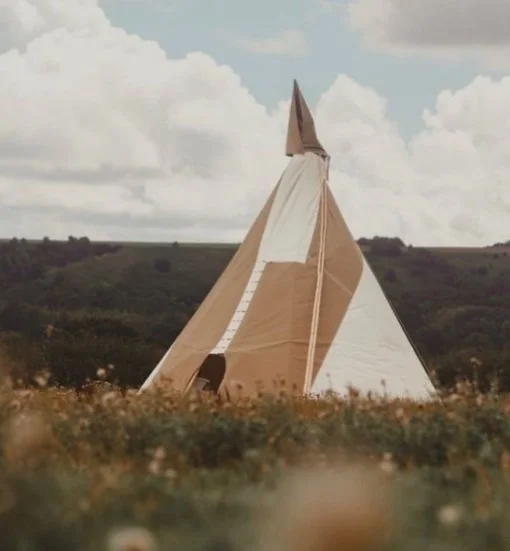 A large teepee-style tent with beige and white fabric panels set up in an open field, with rolling hills and a cloudy sky in the background.