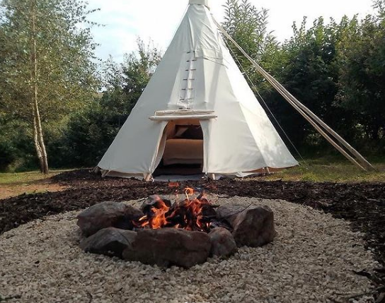 A Native American-style teepee tent set up outdoors with a campfire surrounded by rocks in the foreground.