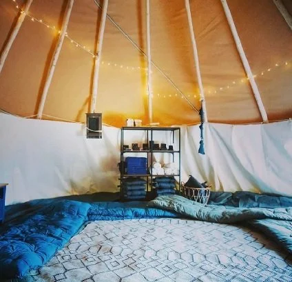 Interior of a teepee tent with string lights, a black shelf holding toiletries and towels, and a bed with blue and white bedding.