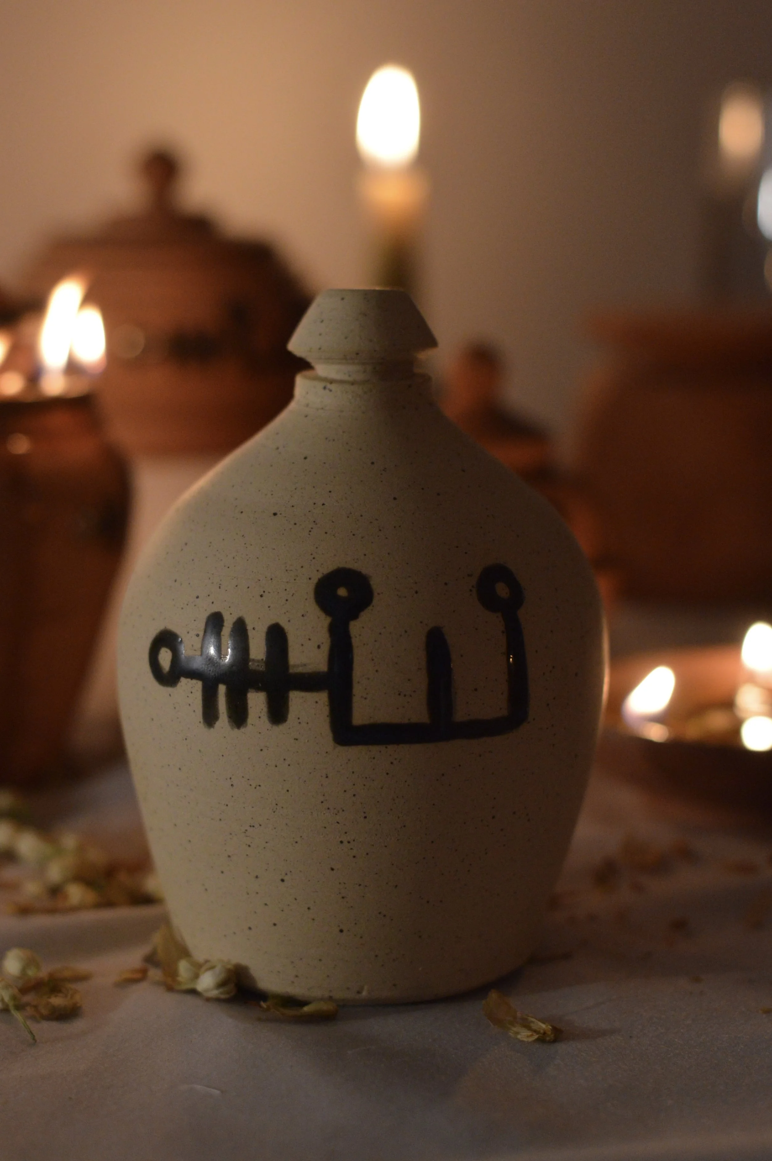 A beige ceramic vase with a black abstract line drawing on it, set against a background of dimly lit candles and pottery. The surface is speckled, and the scene includes scattered dried flower petals.