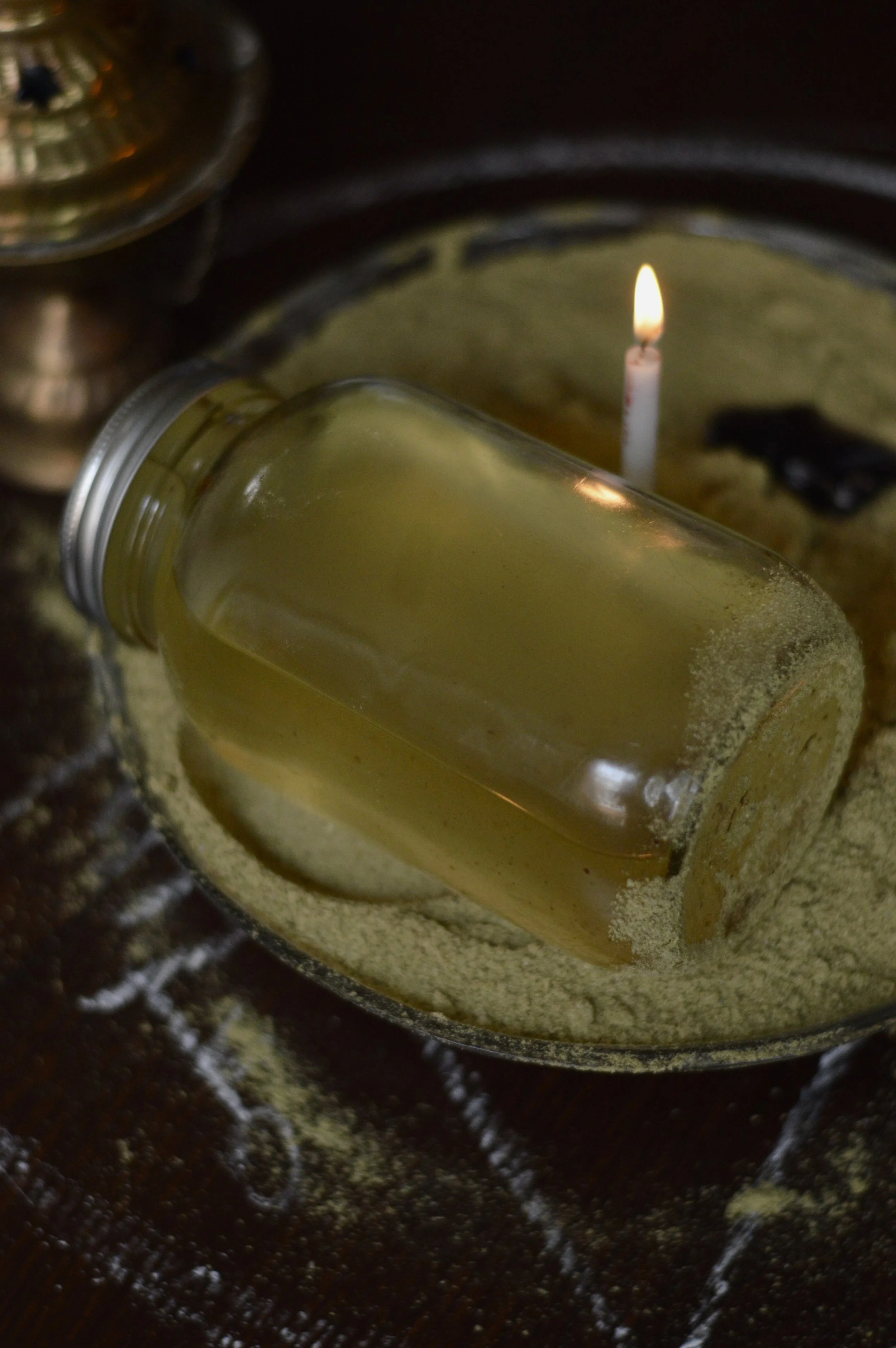 A glass jar filled with a yellowish liquid, partially buried in a mound of powder, on a wooden surface. A lit candle and another jar are visible in the background.